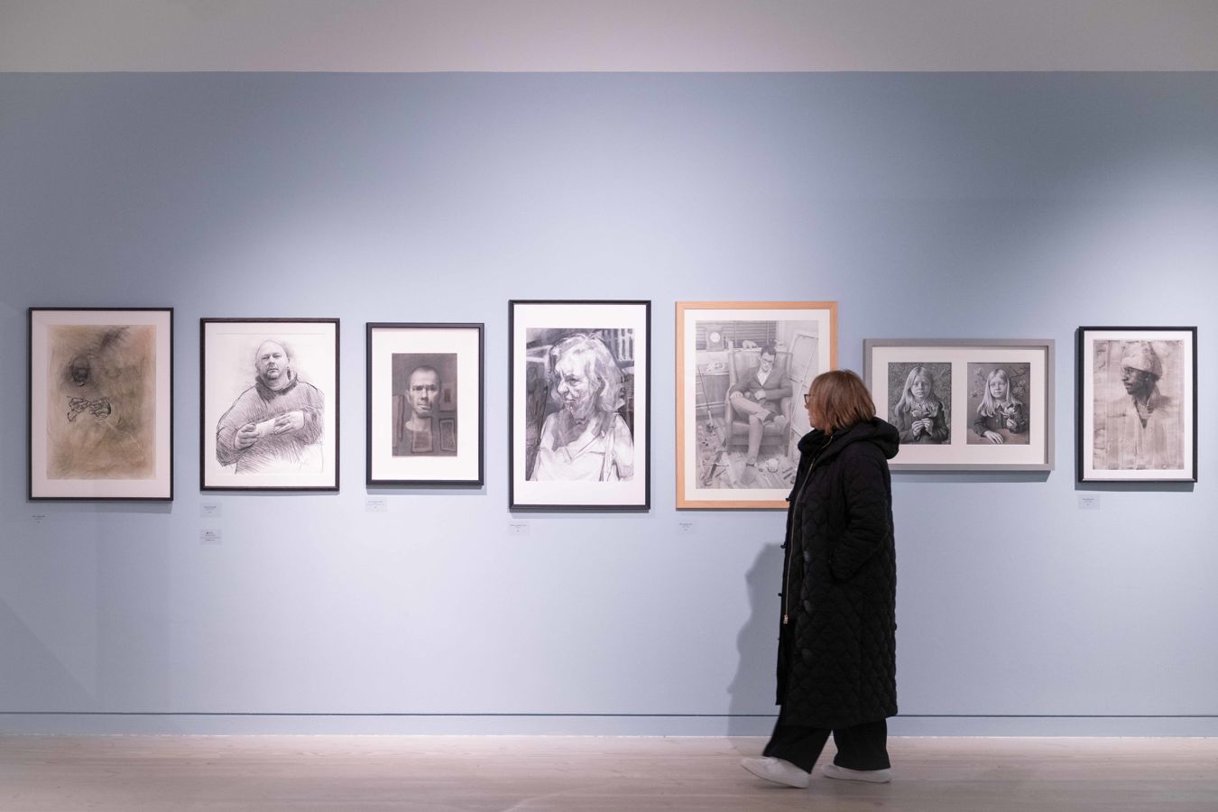 A person in a dark coat observes a gallery wall with seven framed monochrome portraits against a light blue backdrop.