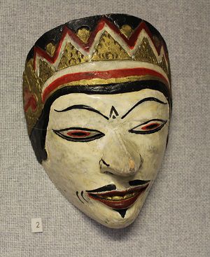 A traditional painted mask with a white face, black eyebrows, and red lips with gold, red, and black patterns.