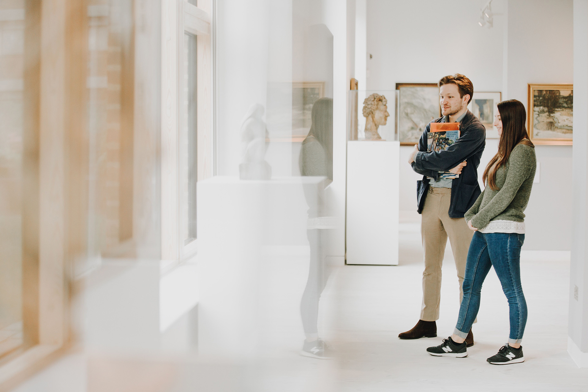 A man and a woman stand in a bright, minimalist art gallery, engaging with the exhibits. 
