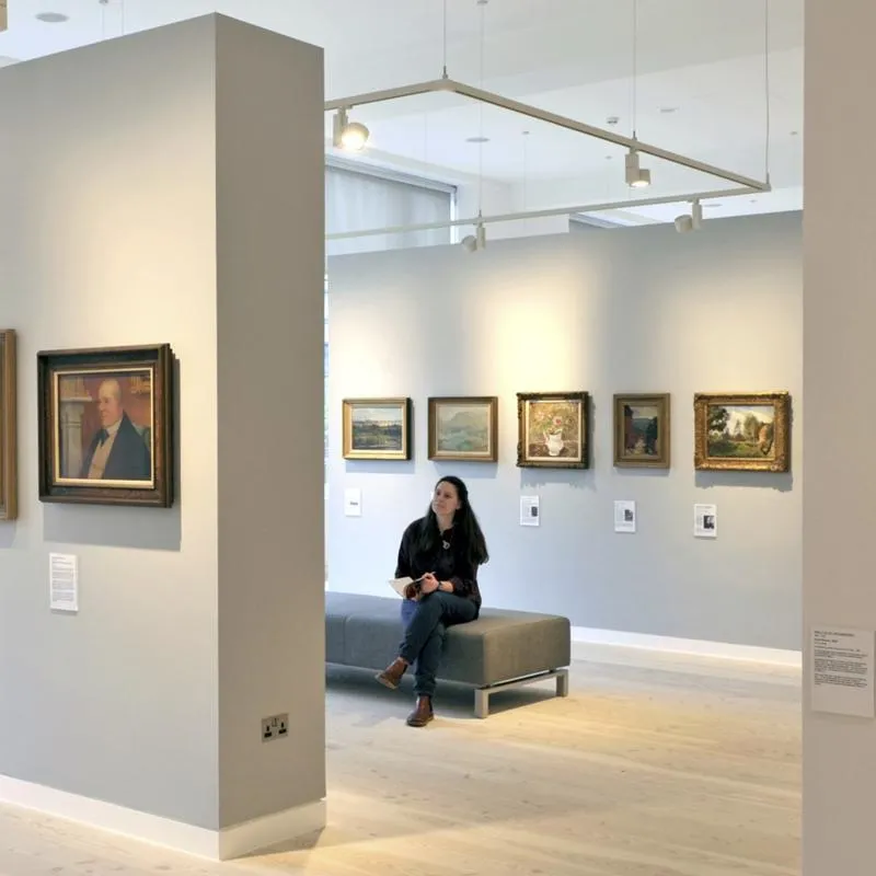 A person sits on a bench in an art gallery with light grey walls, admiring framed paintings.